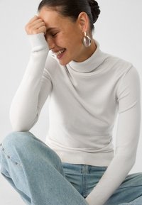 Bela, rebrasta turtleneck pulover z dolgimi rokavi, v kombinaciji z svetlo modrim jeansom. Opazne okrogle uhane dodajajo pridih kontrasta.
