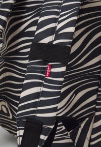 Ryggsäck med svart och krämfärgad zebramönstrad tyg, utrustad med svarta remmar och en liten röd Levi's-logotyp. Texturerad yta med konturerad design.