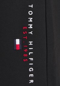 Tela negra con texto bordado en blanco y rojo que dice "TOMMY HILFIGER EST. 1985," con un pequeño bloque de logo tricolor.