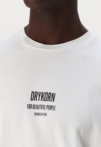 Valge puuvillane T-särk, millel on musta värviga trükitud tekst "DRYKORN FOR BEAUTIFUL PEOPLE". Ümar kaelus ja sile tekstuur.