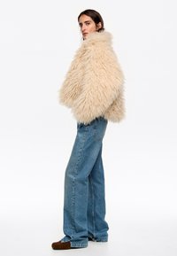 Chaqueta de piel sintética beige con un alto collar, de corte oversized; combinada con jeans de pierna ancha azul claro y zapatos de ante marrón.