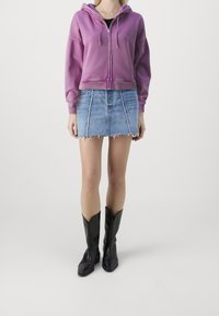 Sudadera con cremallera de color fucsia y capucha con cordón, combinada con una mini falda vaquera con dobladillo sin rematar y costuras verticales, y botas de vaquero negras.