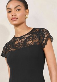 Abito nero con maniche a cappuccio in pizzo con motivi intricati, tessuto morbido e silhouette aderente. Orecchini d'oro completano il look.