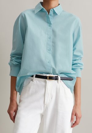 Personne portant une chemise bleu clair boutonnée rentrée dans un pantalon blanc avec une ceinture noire, debout les bras détendus.