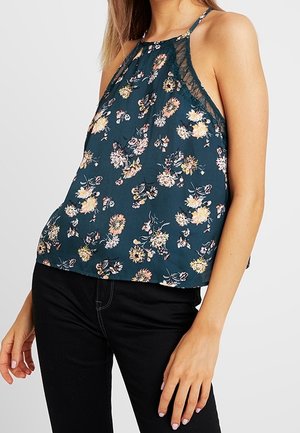 Femme portant un haut sans manches à fleurs bleu sarcelle foncé avec une bordure en dentelle, associé à un pantalon noir ajusté, sur un fond uni.