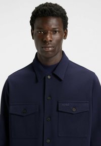 Giacca camicia blu navy realizzata in tessuto testurizzato, con due tasche frontali, chiusure a bottone e un piccolo logo sulla tasca sinistra.