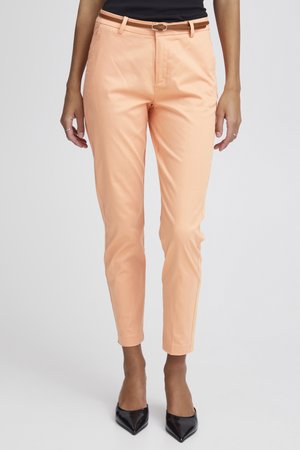 Plānas persiku krāsas bikses ar brūnu jostu, valkātas ar melniem smailiem augstpapēžu kurpēm, pret vienveidīgu gaišu fona.