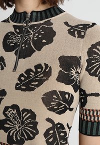 Top de punto con base beige y patrones florales y de hojas en negro, cremallera de un cuarto de longitud y cuello acanalado con acentos en verde y naranja.