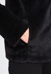 Czarna kurtka z sztucznego futra o miękkiej, teksturowanej powierzchni; posiada boczną kieszeń oraz szerokie rękawy, które podkreślają puszystość materiału.