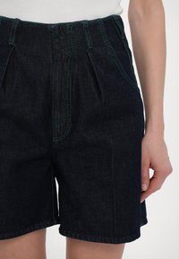 Shorts en denim foncé avec des plis à l'avant, des coutures contrastantes vertes et une coupe décontractée. La ceinture est dotée d'une fermeture à bouton.