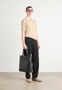 Μπεζ πόλο μπλούζα με κρεμ γιακά, μαύρο ραμμένο παντελόνι και μαύρη δερμάτινη τσάντα. Μοντέλο με σκούρα γυαλιά ηλίου και μπεζ παπούτσια.