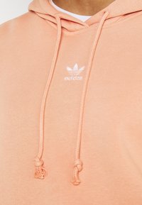 Broskvově zbarvená mikina s kapucí a šňůrkami, s malým bílým logem Adidas. Vyrobena z měkké, texturované látky, s ležérním designem.