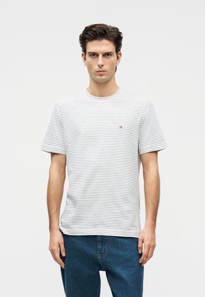 Νέος άνδρας με κοντά σκούρα μαλλιά, φορώντας λευκό ριγέ T-shirt και μπλε τζιν, στέκεται μπροστά σε ένα απλό φωτεινό φόντο.