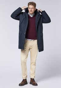 Navy blue padded parka with a hood, featuring two front pockets, worn over a burgundy sweater and beige trousers; brown lace-up boots.