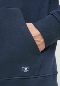 Sudadera azul marino con un bolsillo canguro, puños acanalados y una etiqueta de tela que detalla el nombre de la marca y el año de fundación. Textura suave.