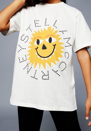 Barn iført hvid T-shirt med et stort gult smilende solansigt og ordet "TELLA" i prikkede bogstaver omkring det.