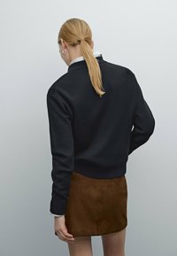 Svart tröja med ribbade ärmar och nederkant, i kombination med en brun mockaskirt. Släta texturer med minimalistisk design. Håret i en hästsvans.