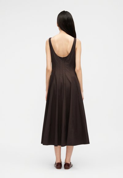 Γυναίκα με μακριά σκούρα μαλλιά που φοράει μίντι φόρεμα σε σκούρο καφέ χρώμα, αμάνικο, με χαμηλή πλάτη και καφέ επίπεδα παπούτσια, στέκεται μπροστά σε απλό φόντο.