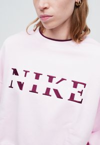 Ružová mikina s okrúhlym výstrihom, na ktorej je tmavočervenou vyšívkou nápis "NIKE" a pod ním v bielej farbe nápis "SPORTSWEAR". Mäkká textúra.