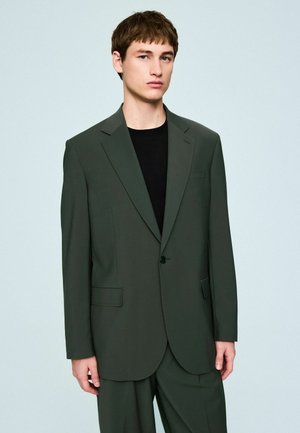 Young man wearing dark green oversized suit jacket and matching trousers over a black shirt, standing against a light blue background.