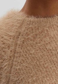 Gros plan d'une personne portant un pull beige doux et duveteux avec un col rond côtelé.