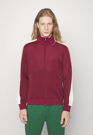 Burgundy zip-up athletic jacket with a high collar, white side panels, and two zippered pockets. Smooth fabric and ribbed cuffs.