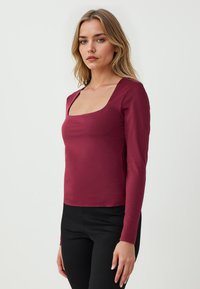 Maglietta rossa a maniche lunghe con scollatura quadrata, realizzata in un tessuto liscio ed elastico. Design aderente con cuciture minime. Abbinata a pantaloni neri.