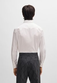 White long-sleeve shirt with a classic collar, paired with dark gray patterned trousers featuring a subtle check design and button accents.