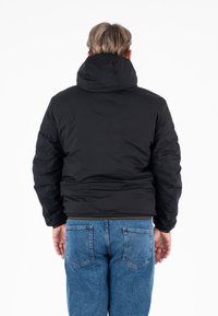 Giacca nera imbottita con cappuccio, caratterizzata da una texture liscia e polsini elasticizzati. Indossata con jeans in denim blu. Vista posteriore mostrata.