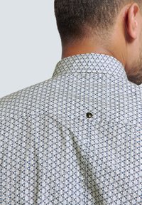 Hemd mit Knöpfen aus einem hellen Stoff mit geometrischem Muster in Marineblau und Rot, ausgestattet mit einem klassischen Kragen und einem Knopfdetail auf dem Rücken.