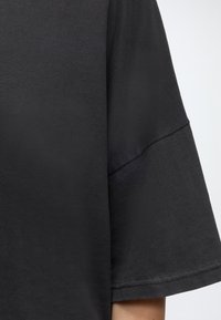 Gros plan d'une personne portant une chemise ample à manches courtes gris foncé avec des détails visibles de couture sur la manche.