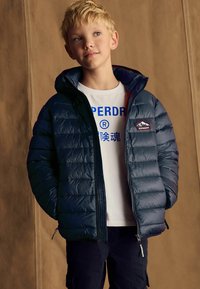 Chaqueta acolchada marina con capucha desmontable, con cierre de cremallera y un parche con el logo. Llevada sobre una camiseta blanca con estampado azul.