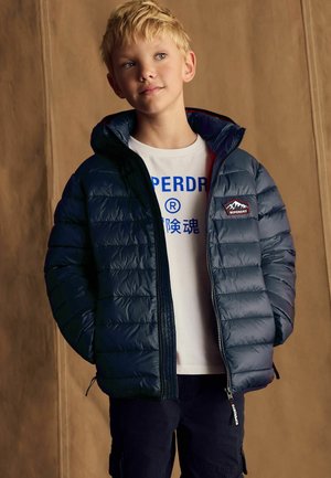 Chaqueta acolchada marina con capucha desmontable, con cierre de cremallera y un parche con el logo. Llevada sobre una camiseta blanca con estampado azul.