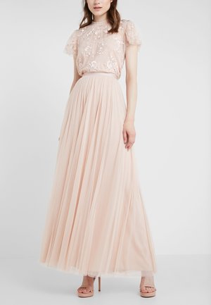 Femme portant un chemisier transparent rose poudré brodé de fleurs, associé à une jupe longue plissée en tulle taille haute et des talons nude.