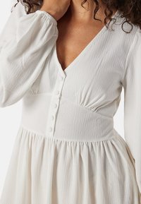 Abito bianco realizzato in un tessuto leggero e testurizzato. Presenta un collo a V, dettagli con bottoni e vita arricciata, che sfocia in una gonna aderente.