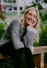 Hallide ja valgete triipudega kudum, millel on relaxed fit, soonikstruktuur ja laiad mansetid. Tumedad püksid. Istub puidust pingil rohelises keskkonnas.