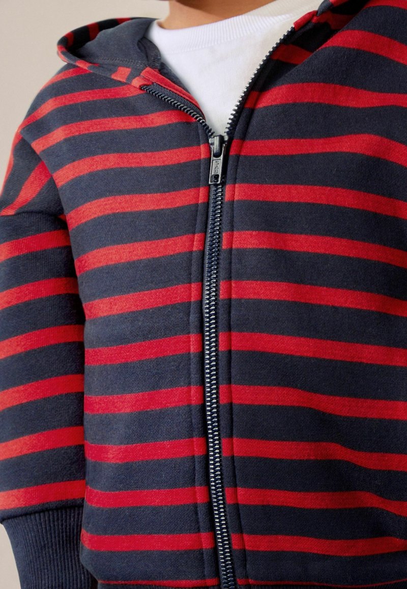 Enfant portant un sweat à capuche zippé à rayures bleu marine et rouge sur un t-shirt blanc, torse visible, fermeture éclair partiellement fermée.