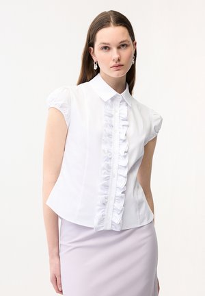 Ung kvinde med langt brunt hår iført en hvid kortærmet bluse med flæser ned foran og en lyse lilla nederdel.