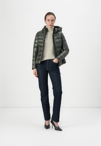 Doudoune verte avec capuche, finition texturée, fermeture éclair à l'avant, portée avec un pull en tricot gris en dessous, associée à un jean foncé et des talons noirs.