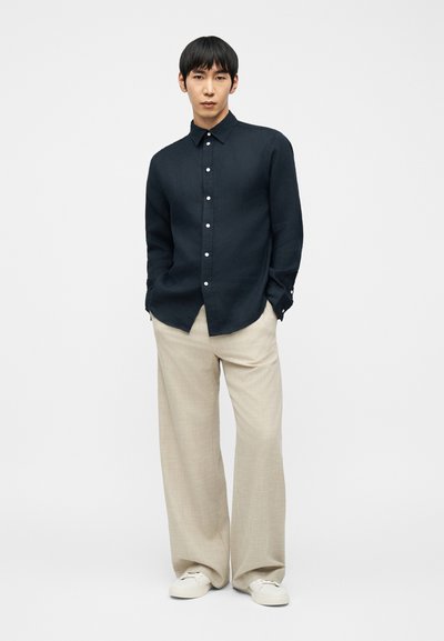 Νέος άνδρας που στέκεται με τα χέρια στις τσέπες, φορώντας μαύρο πουκάμισο με κουμπιά, μπεζ παντελόνι φαρδιάς γραμμής και λευκά αθλητικά παπούτσια σε λευκό φόντο.