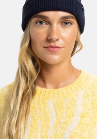 Geel gebreid sweater met een textuurpatroon, met een ronde hals. Model draagt een donkere beanie. Achtergrond is helder wit.