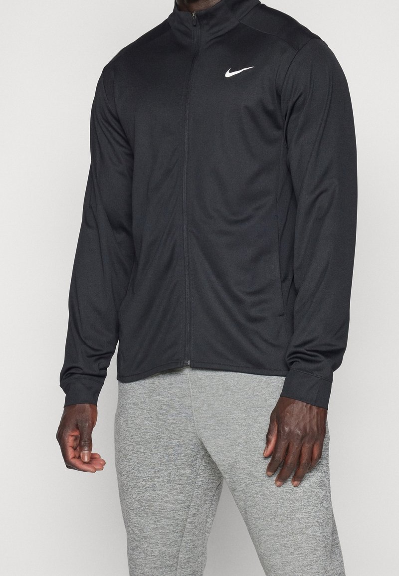 Veste zippée noire Nike à manches longues, en tissu lisse, arborant un logo blanc sur la poitrine, associée à un pantalon de sport gris.