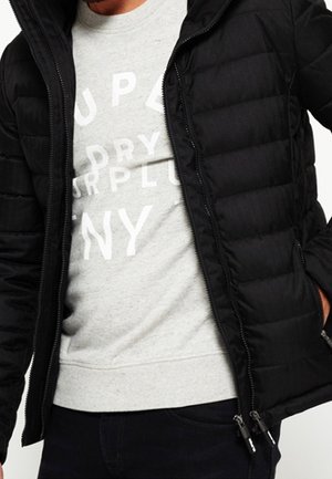 Mężczyzna ubrany w czarną pikowaną kurtkę na jasnoszarej bluzie z białymi nadrukowanymi literami, stojący z jedną ręką w kieszeni.