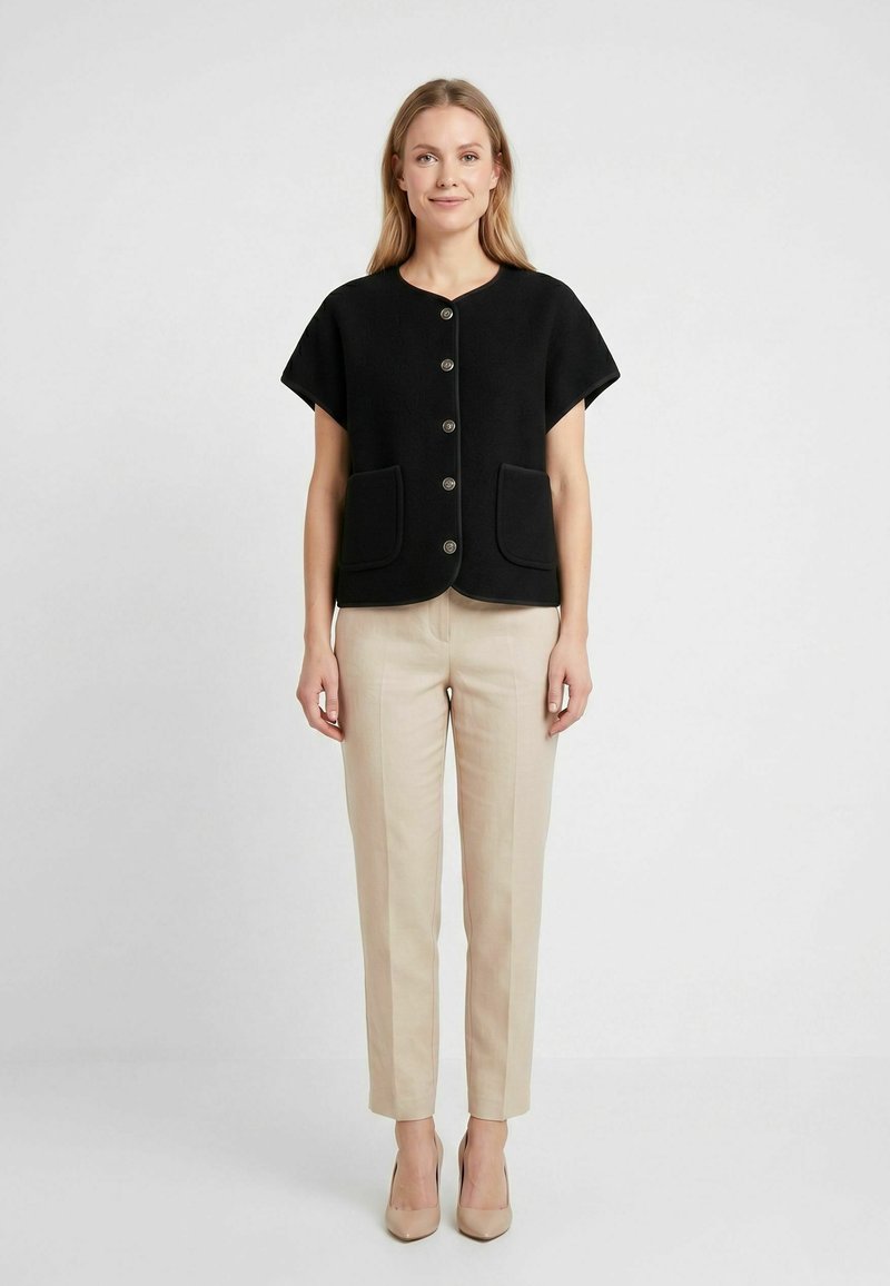 Femme debout sur un fond blanc, portant une veste noire à manches courtes boutonnée, un pantalon beige et des talons hauts beiges.