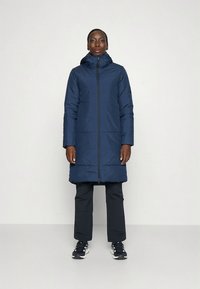 Σκούρο μπλε μπουφάν puffer με φερμουάρ, μακριά μανίκια και κουκούλα. Διαθέτει οριζόντια ραφή και ελαφρώς φαρδιά εφαρμογή. Συνδυάζεται με μαύρο παντελόνι και αθλητικά παπούτσια.