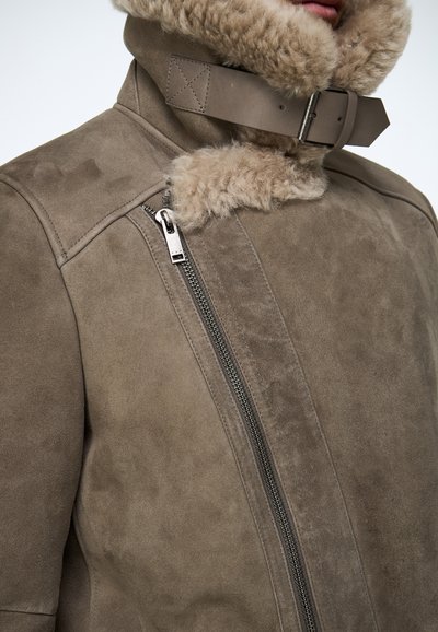 Bruine suède jas met een pluizige beige kraag, een zijrits en een gespband bij de nek. Zachte textuur en minimalistisch ontwerp.