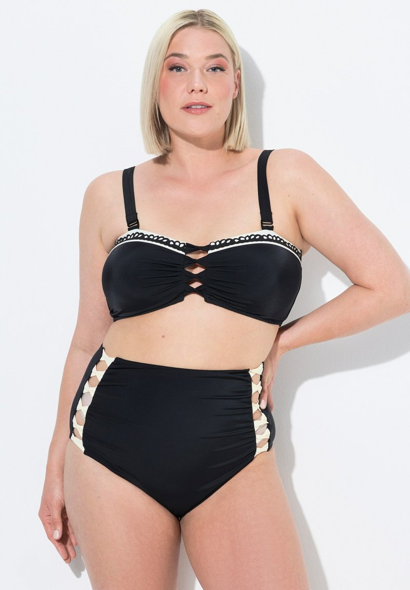 Černý bikiny top s nařaseným materiálem, křížovým detailem vpředu a bílým zoubkovaným lemem; v kombinaci s černými vysokými kalhotkami s bočními výřezy.