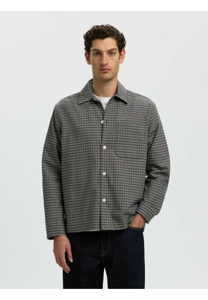 Homme portant une chemise à carreaux grise boutonnée sur un t-shirt blanc avec une main dans la poche d'un jean foncé sur un fond uni.