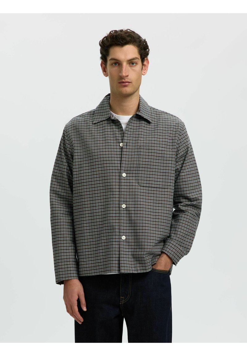 Homme portant une chemise à carreaux grise boutonnée sur un t-shirt blanc avec une main dans la poche d'un jean foncé sur un fond uni.