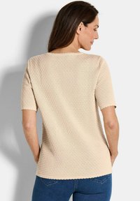 Eine Frau mit langen braunen Haaren, die ein beiges, strukturiertes kurzärmliges Oberteil und blaue Jeans trägt, steht mit dem Rücken zu einem hellen Hintergrund.